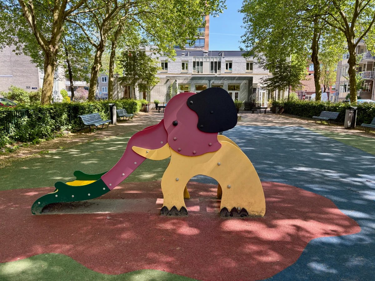 Red and yellow elephant-shaped slide with large shaded plaza and white building behind