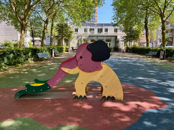 Red and yellow elephant-shaped slide with large shaded plaza and white building behind