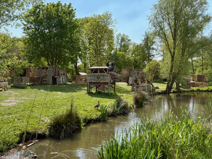 Children sitting on the roof of a hand-built hut on a sunny day with multiple structures set by a canal and trees