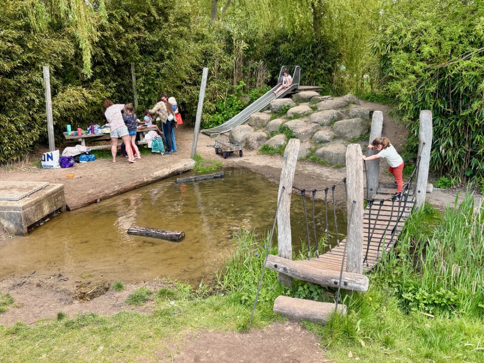 Kids and parents play next to a small pond with wooden bridge, slide, and rocks