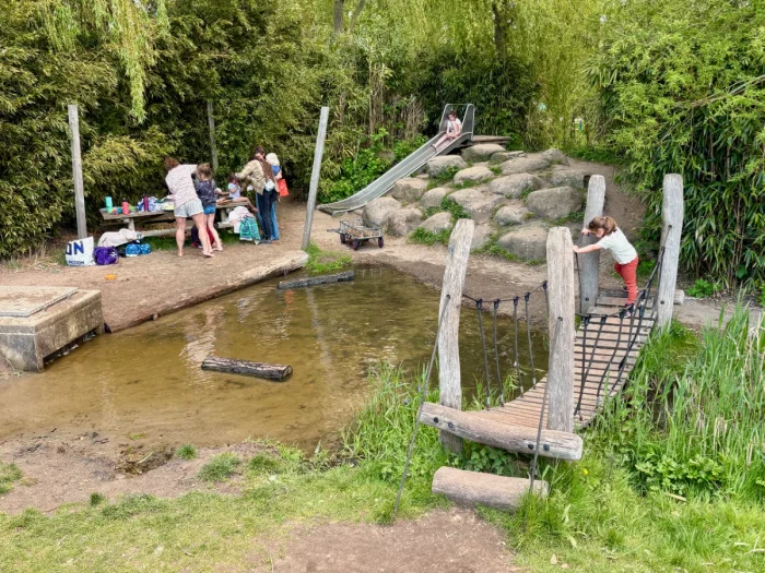 Kids and parents play next to a small pond with wooden bridge, slide, and rocks