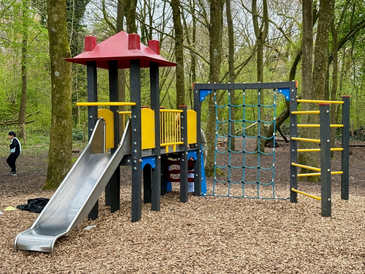 Red, blue, and yellow children's play structure with slide, climbing net, and ladder surrounded by forest