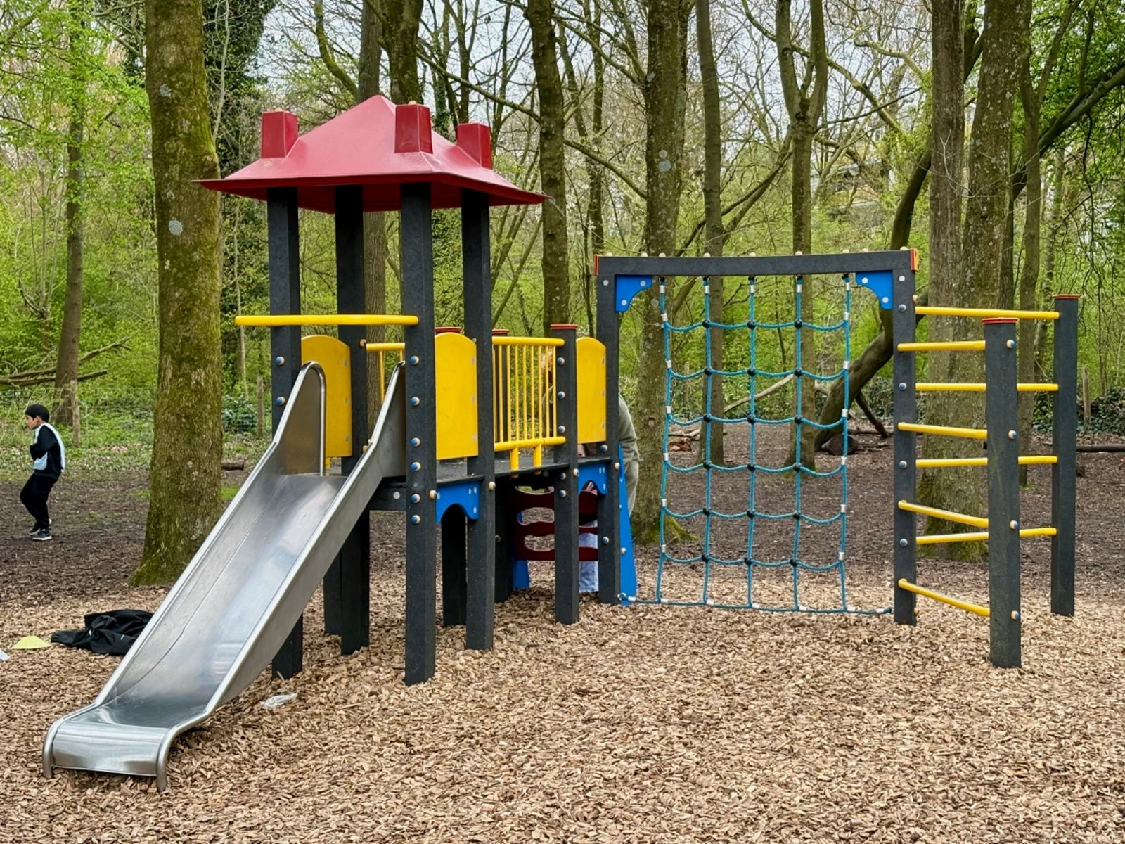 Red, blue, and yellow children's play structure with slide, climbing net, and ladder surrounded by forest