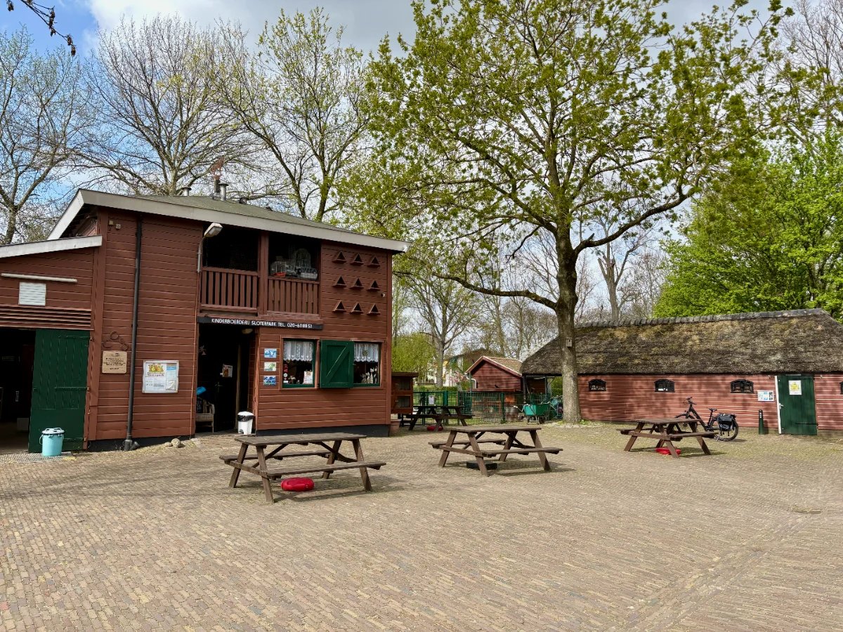 Brown farm building with green door and shutters with brick terrace, picnic tables, and mature trees