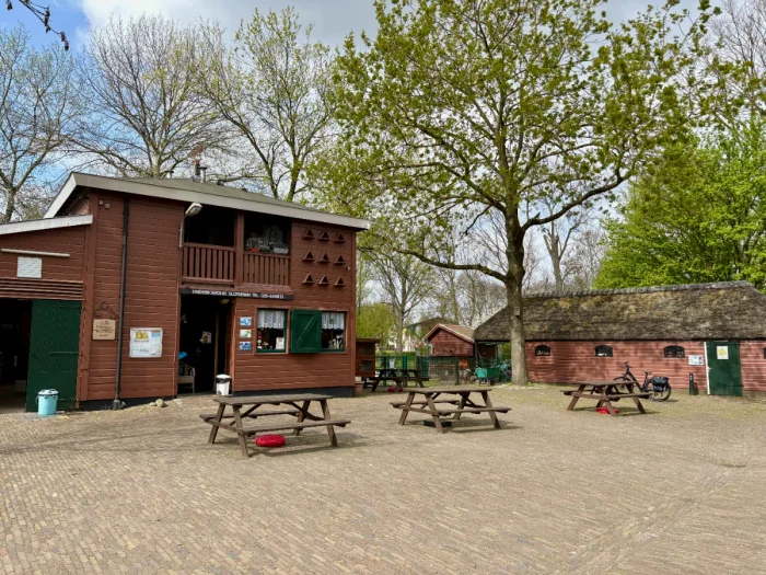 Brown farm building with green door and shutters with brick terrace, picnic tables, and mature trees