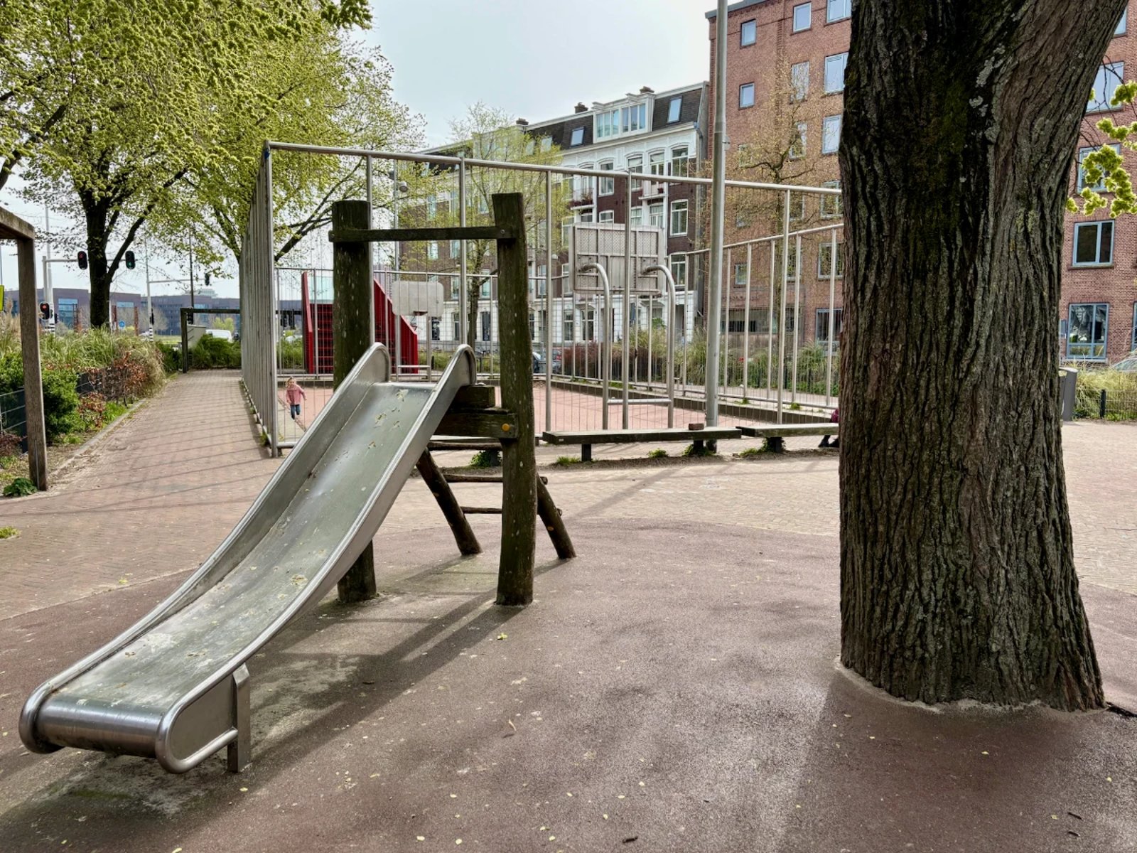 Slide with wooden ladder and kid-sized enclosed basketball court and large tree adjacent