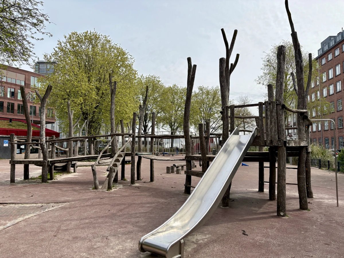 Natural wooden playground structure with bridge, ladder, and slide and large trees behind