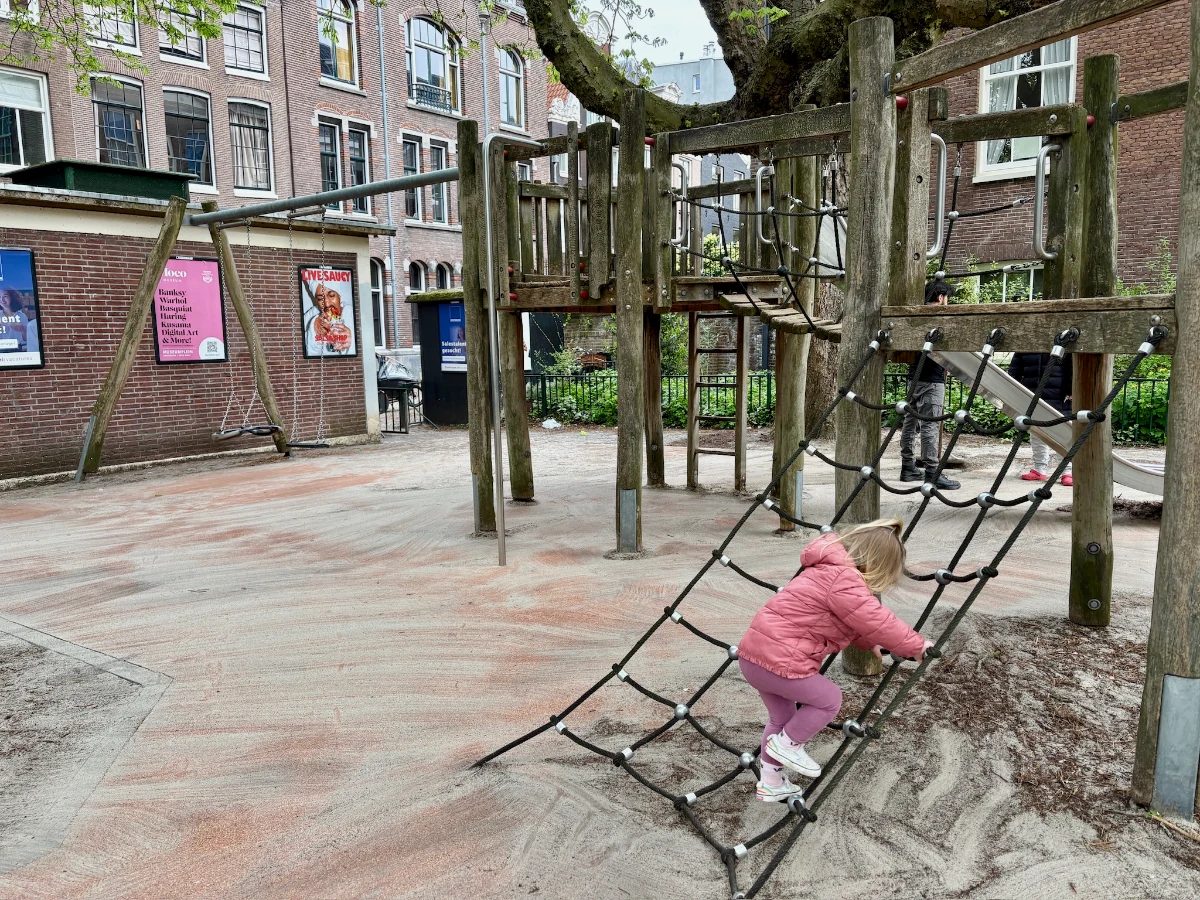 Girl in pink coat on climbing net leading to wooden play structure next to brick building with colorful posters