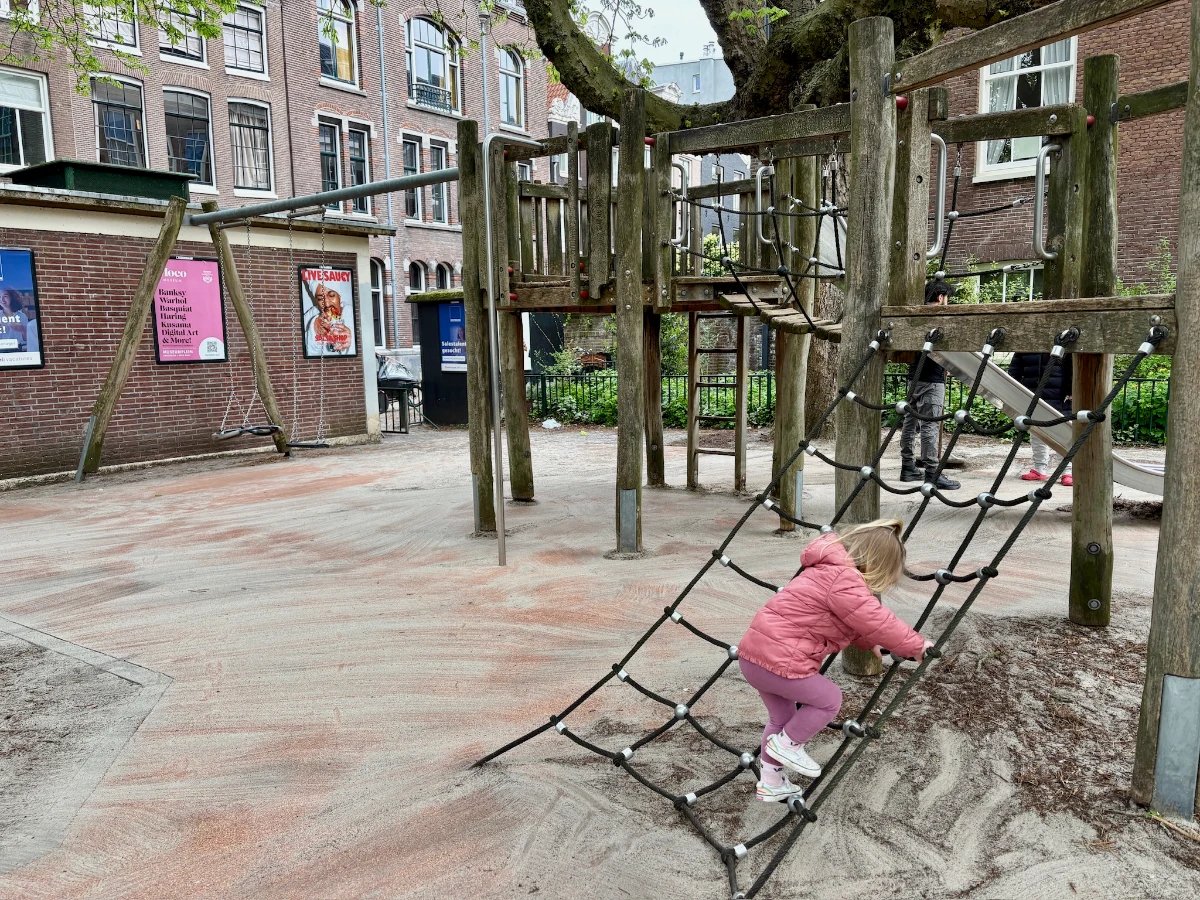 Girl in pink coat on climbing net leading to wooden play structure next to brick building with colorful posters