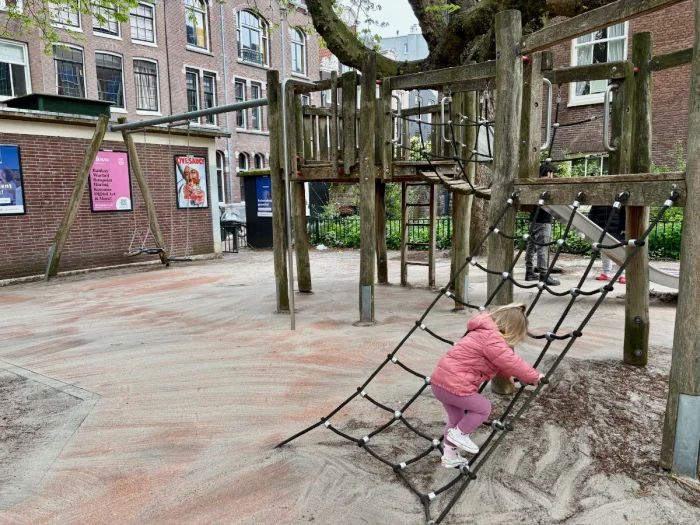 Girl in pink coat on climbing net leading to wooden play structure next to brick building with colorful posters