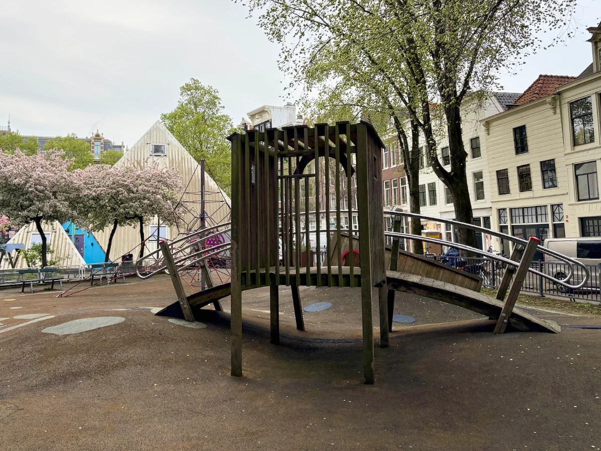 Arched wooden bridge on playground with modern pyramid-shaped building, spring trees, and houses behind