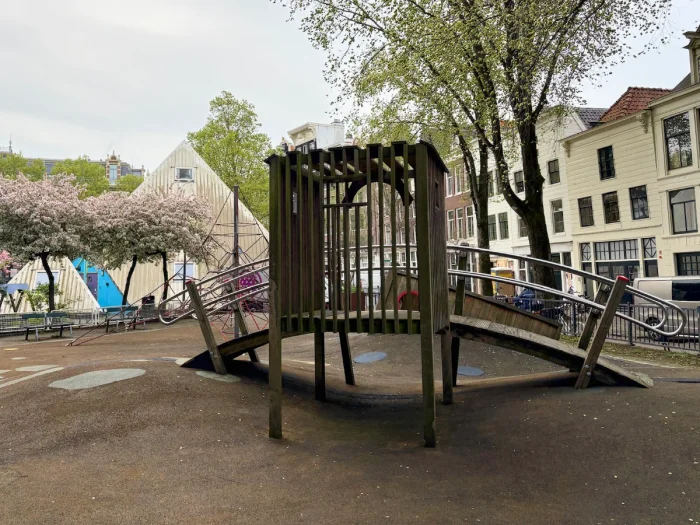 Arched wooden bridge on playground with modern pyramid-shaped building, spring trees, and houses behind
