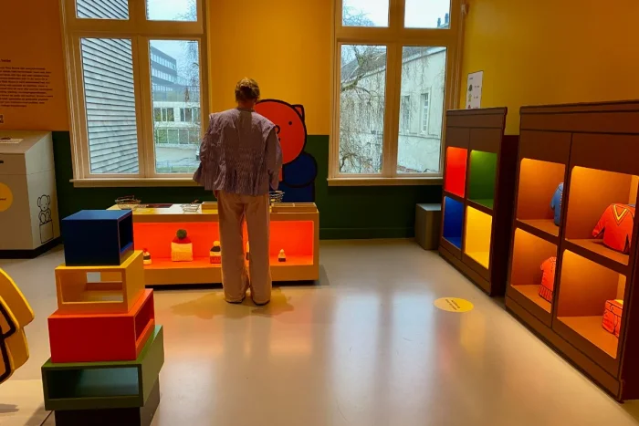 Parent standing in a play room designed like a shop with primary colors, shelves with clothing, and a checkout counter