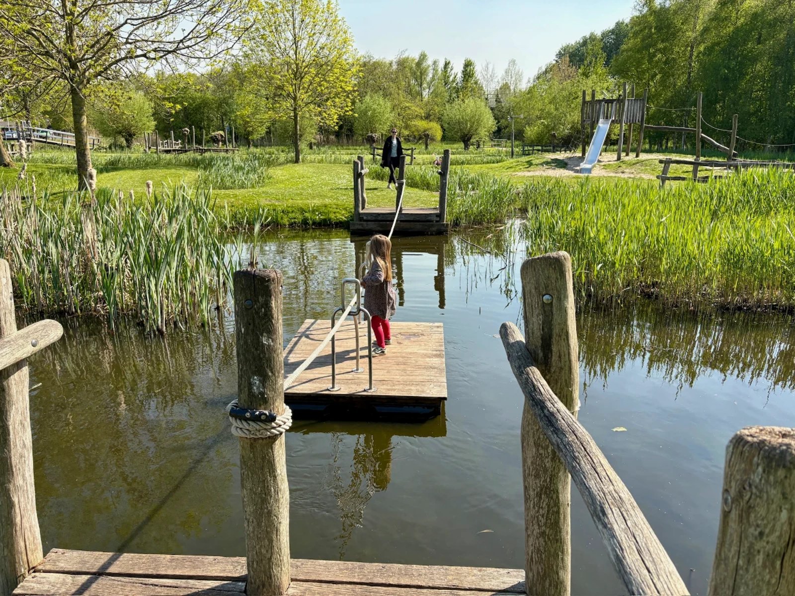 Young girl using hand-operated ferry moving across a canal with tall grass and sunny lawn on other side