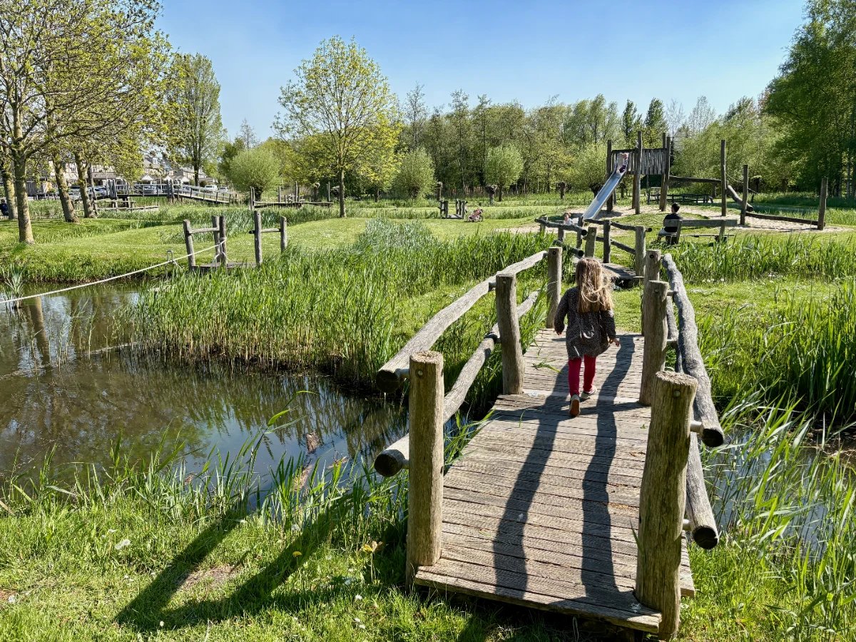 Girl walking across rustic wooden bridge over canal with playground on other side