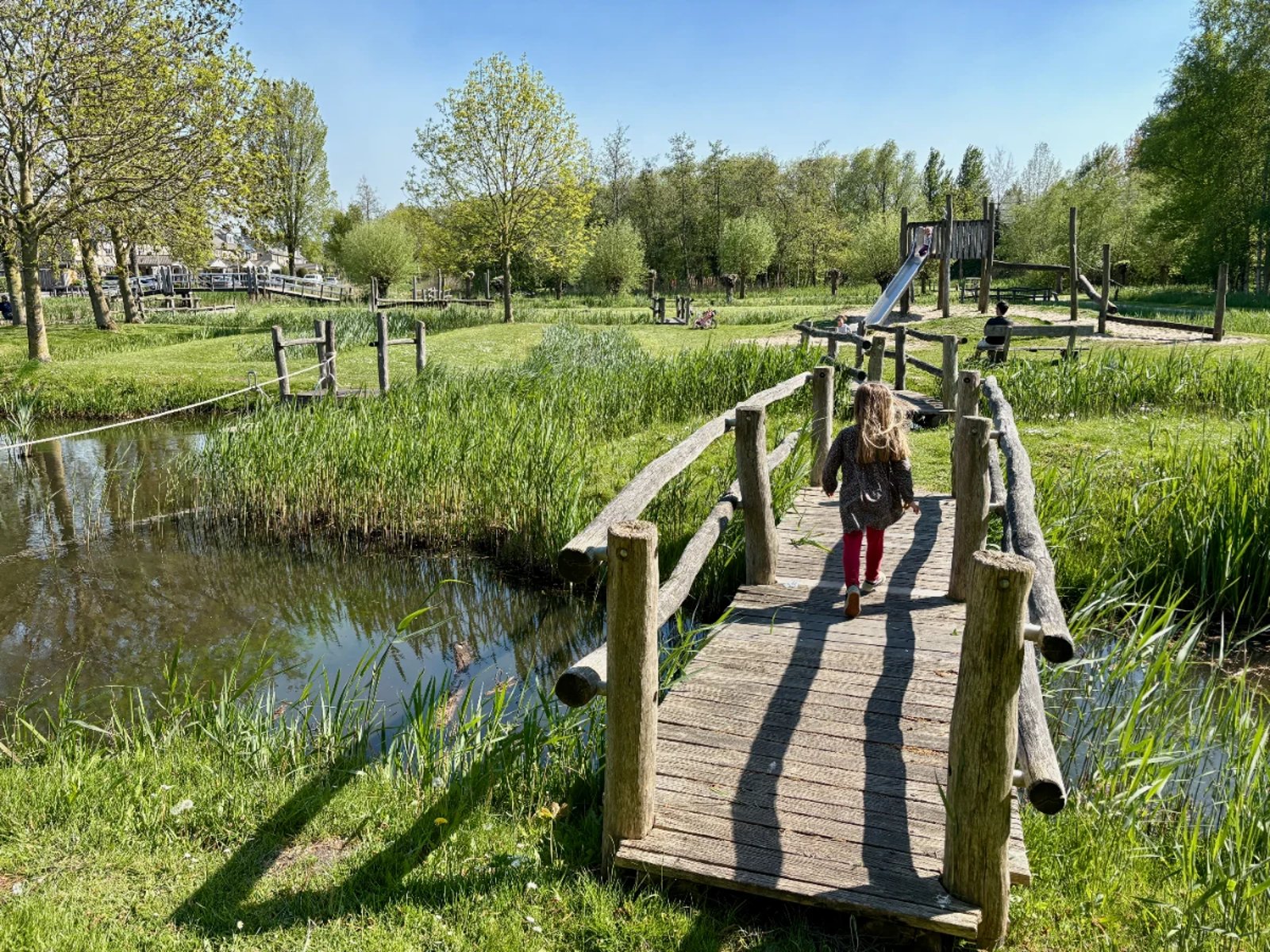 Girl walking across rustic wooden bridge over canal with playground on other side