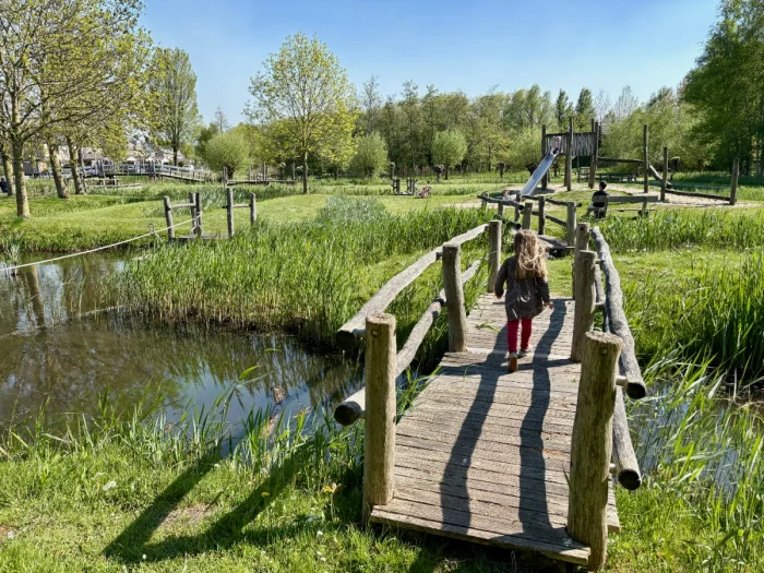 Girl walking across rustic wooden bridge over canal with playground on other side