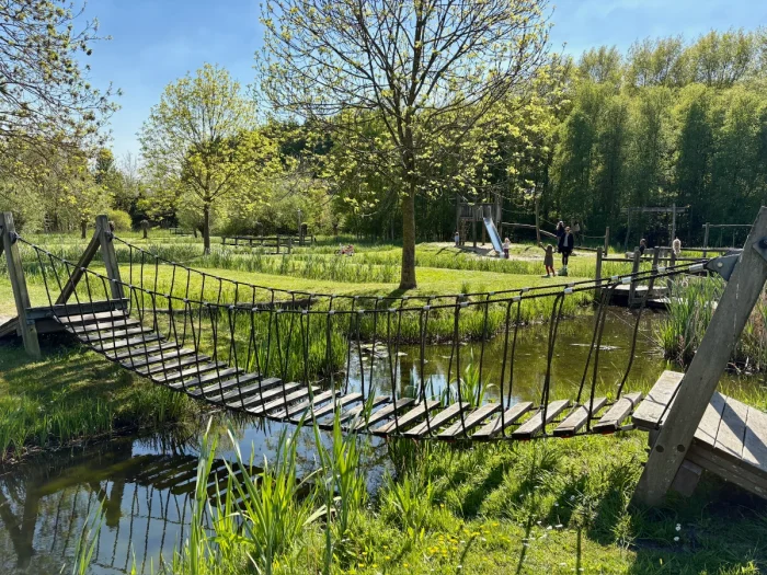 Wooden slat bridge with ropes and green lawn with trees and playground behind