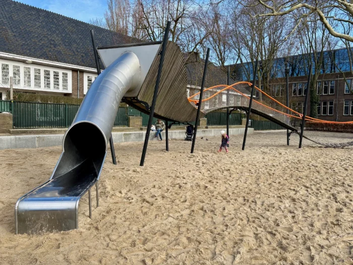 Large enclosed metal slide connected to wooden play structure and wavy walkway with sand below