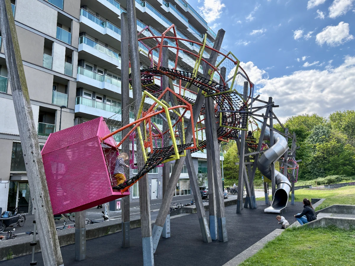 Modern pink, yellow, and orange play garland elevated on wooden stilts with tall curved slide next to modern apartment building