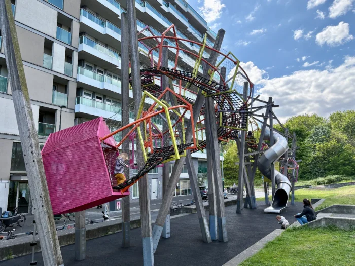 Modern pink, yellow, and orange play garland elevated on wooden stilts with tall curved slide next to modern apartment building
