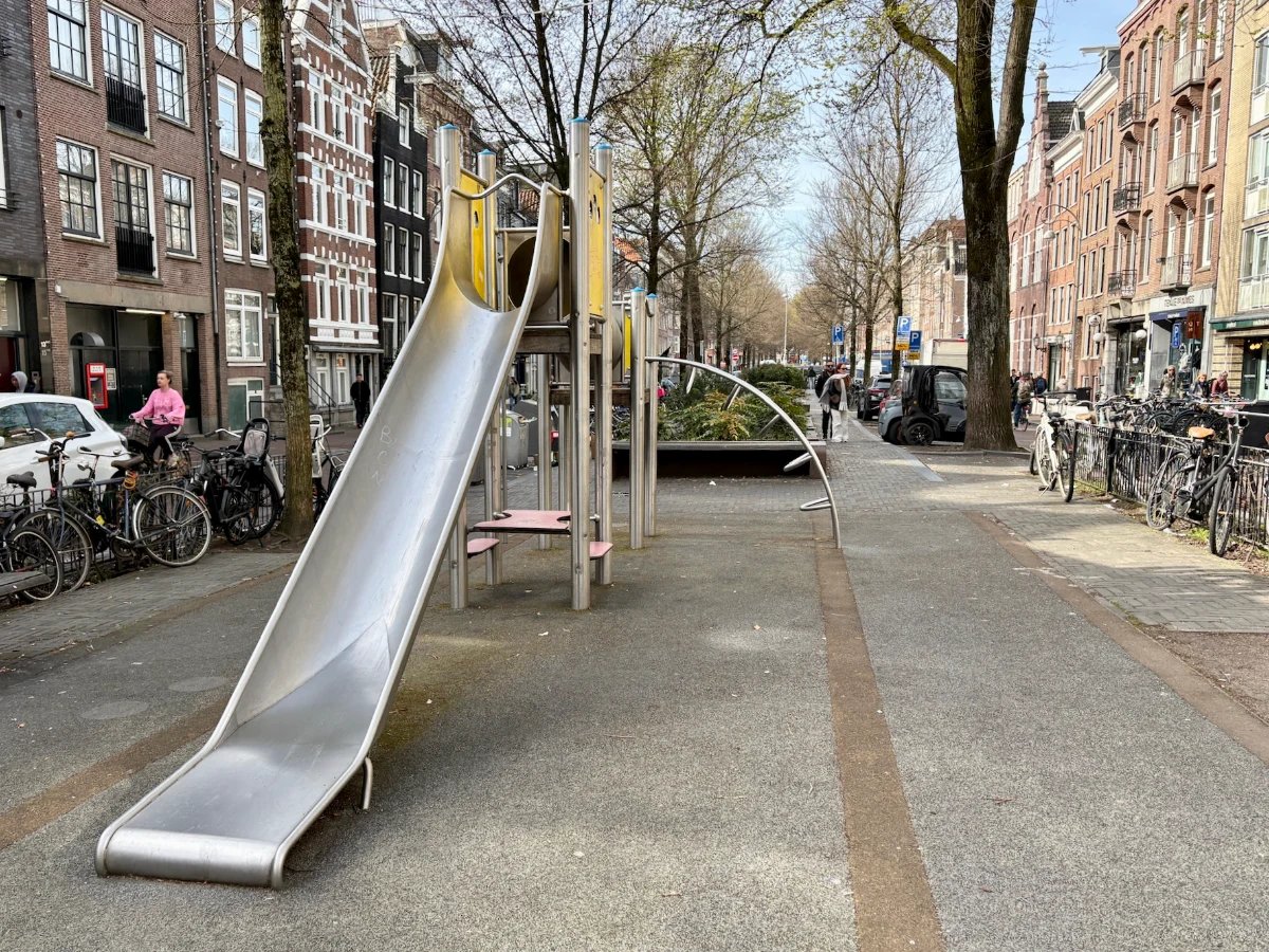 Metal climbing structure with slide and arched ladder set within a city block with buildings, bikes, cars on either side