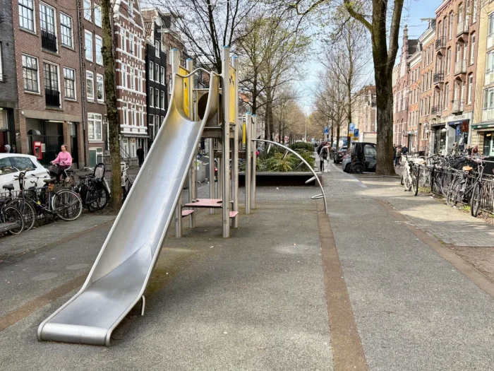 Metal climbing structure with slide and arched ladder set within a city block with buildings, bikes, cars on either side