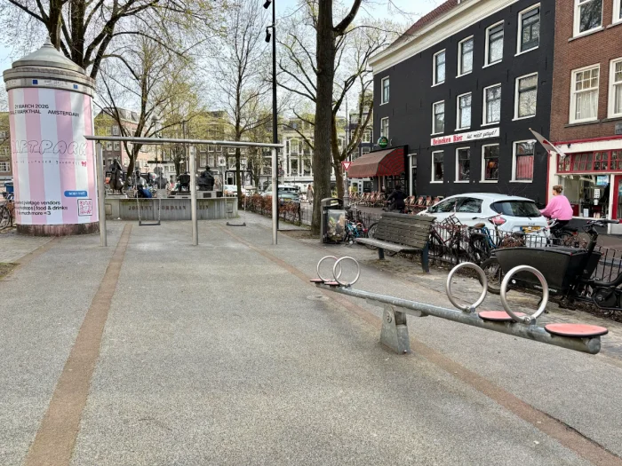 Metal seesaw and swing set on concrete with buildings, cars, and a pink and white striped advertisement on a peperbus