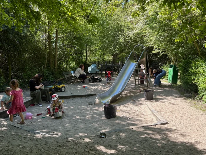 Kids and parents playing on small sandy and shaded playground with slide, toys and benches