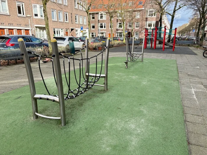 Playground with rope obstacle course and red climbing structure behind with parked cars and residential buildings surrounding