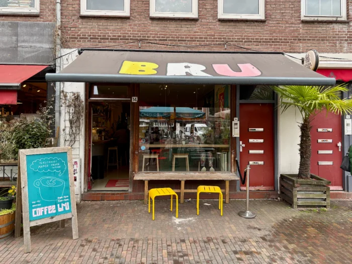 Coffee shop exteior with bench and bright yellow stools, blue sandwich board, and brown awning with BRU written on it