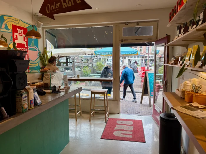 Looking toward entrance of coffee shop with bar, shelving with merchandise, colorful artwork and a large window with bar seating