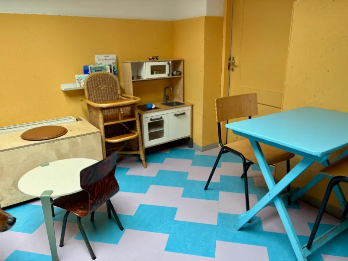 Play kitchen and high chair next to blue table and chairs in room with yellow walls and blue and purple checkered floor