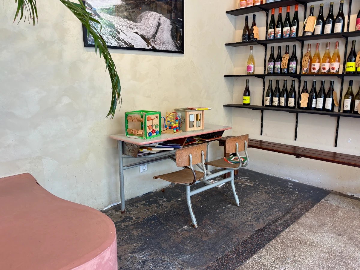 Small children's desk with two chairs and toys on top next to a palm and shelves with wine bottles