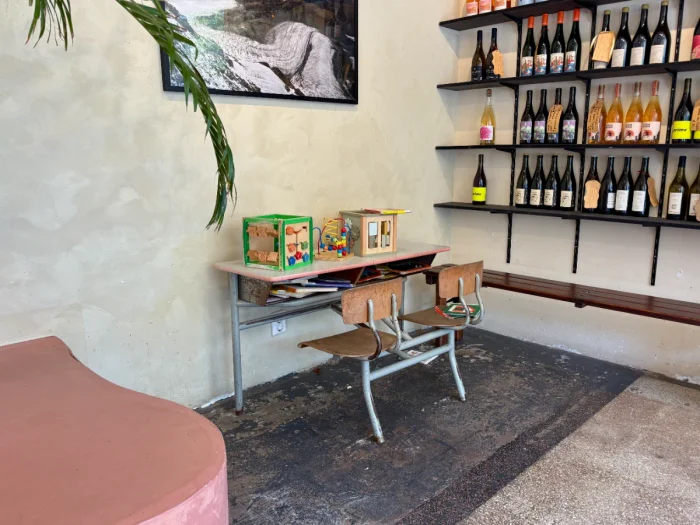 Small children's desk with two chairs and toys on top next to a palm and shelves with wine bottles
